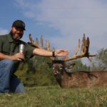 Hunter kneeling with huge whitetail buck kill extending his arm to show size of massive rack