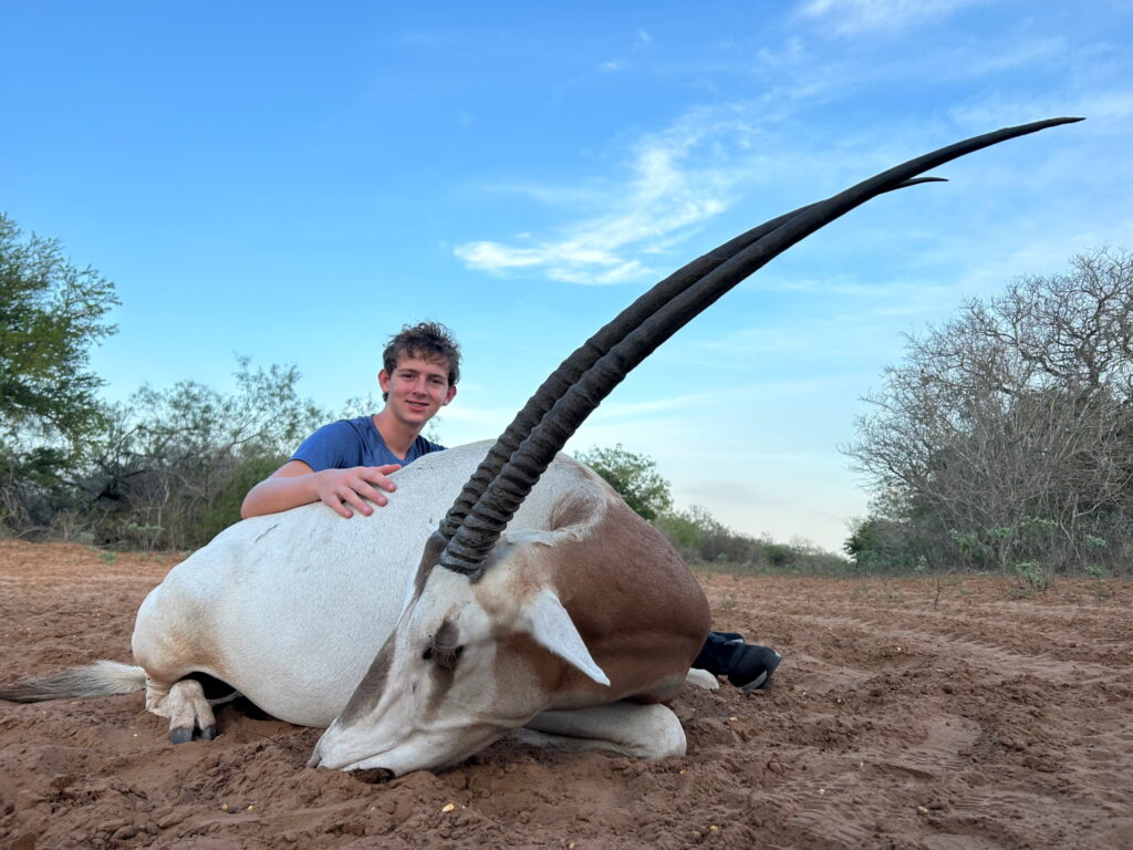 Teenage boy smiling and kneeling beside a Scimitar Horned Oryx lying on the ground at El Monte Gringo Ranch